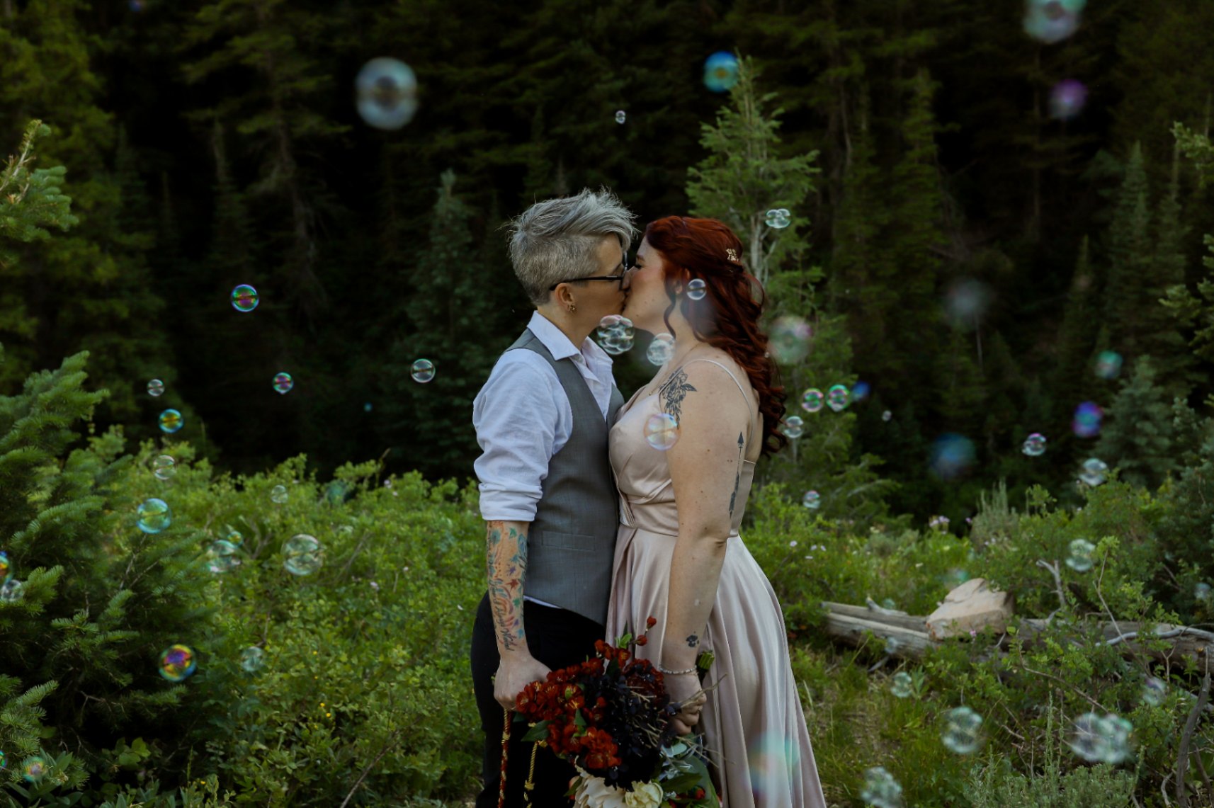Two lesbian brides (both white, one with short gray hair, one with long dark red hair) stand on a mountain. They each have tattoos. The nonbinary bride with short gray hair is wearing a gray vest, a white shirt and dark pants, as well as glasses. The red-haired bride is wearing a light pink dress with spaghetti straps. Bubbles are floating around them.