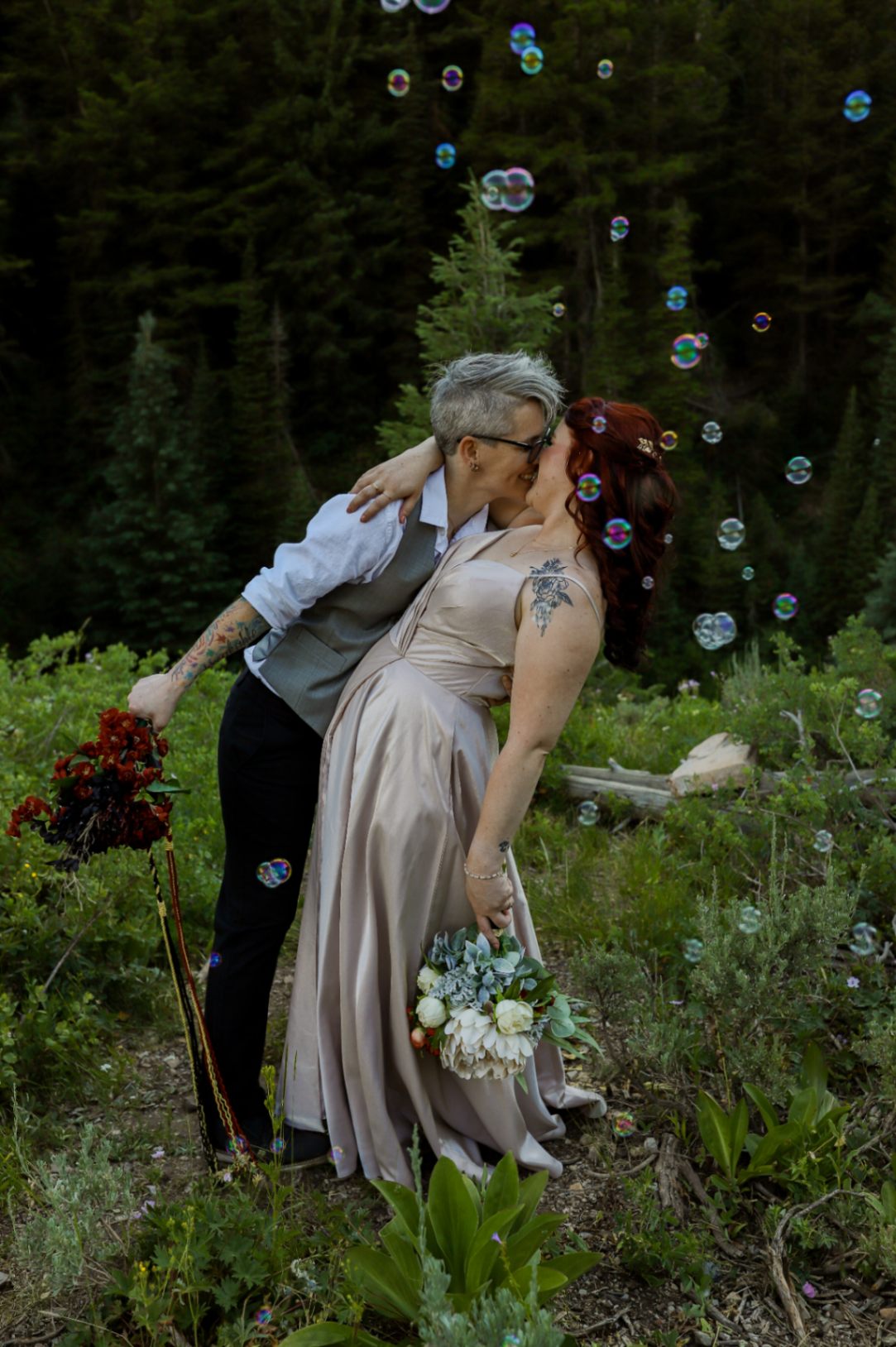 Two lesbian brides (both white, one with short gray hair, one with long dark red hair) kiss with a dip. They each have tattoos. The nonbinary bride with short gray hair is wearing a gray vest, a white shirt and dark pants, as well as glasses. The red-haired bride is wearing a light pink dress with spaghetti straps. The brides are both holding bouquets. Bubbles are floating around them.