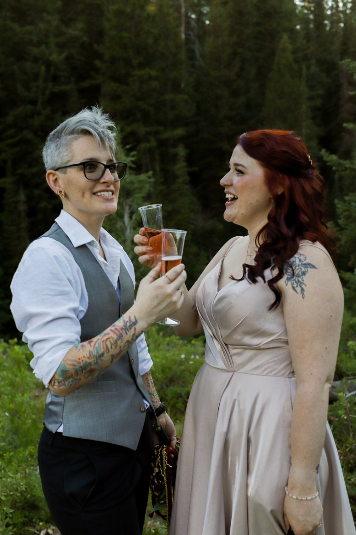 Two lesbian brides (both white, one with short gray hair, one with long dark red hair) toast with sparking rose. They each have tattoos. The nonbinary bride with short gray hair is wearing a gray vest, a white shirt and dark pants, as well as glasses. The red-haired bride is wearing a light pink dress with spaghetti straps.