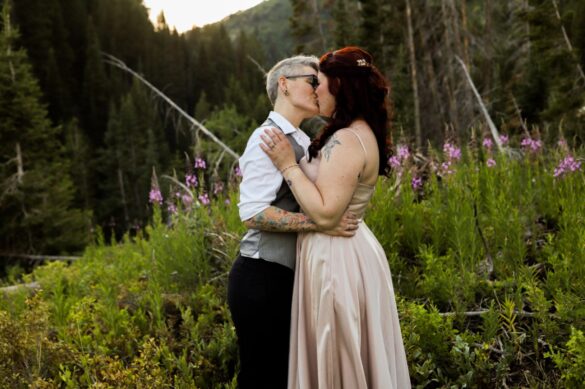 Two lesbian brides (both white, one with short gray hair, one with long dark red hair) kiss. They each have tattoos. The nonbinary bride with short gray hair is wearing a gray vest, a white shirt and dark pants, as well as glasses. The red-haired bride is wearing a light pink dress with spaghetti straps.