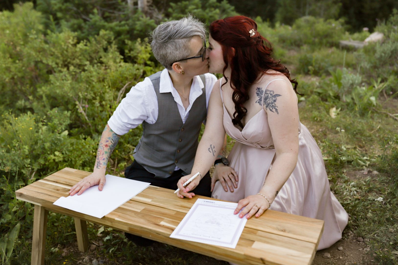 Two brides (both white, one with short gray hair, one with long dark red hair) kiss after signing their marriage certificate