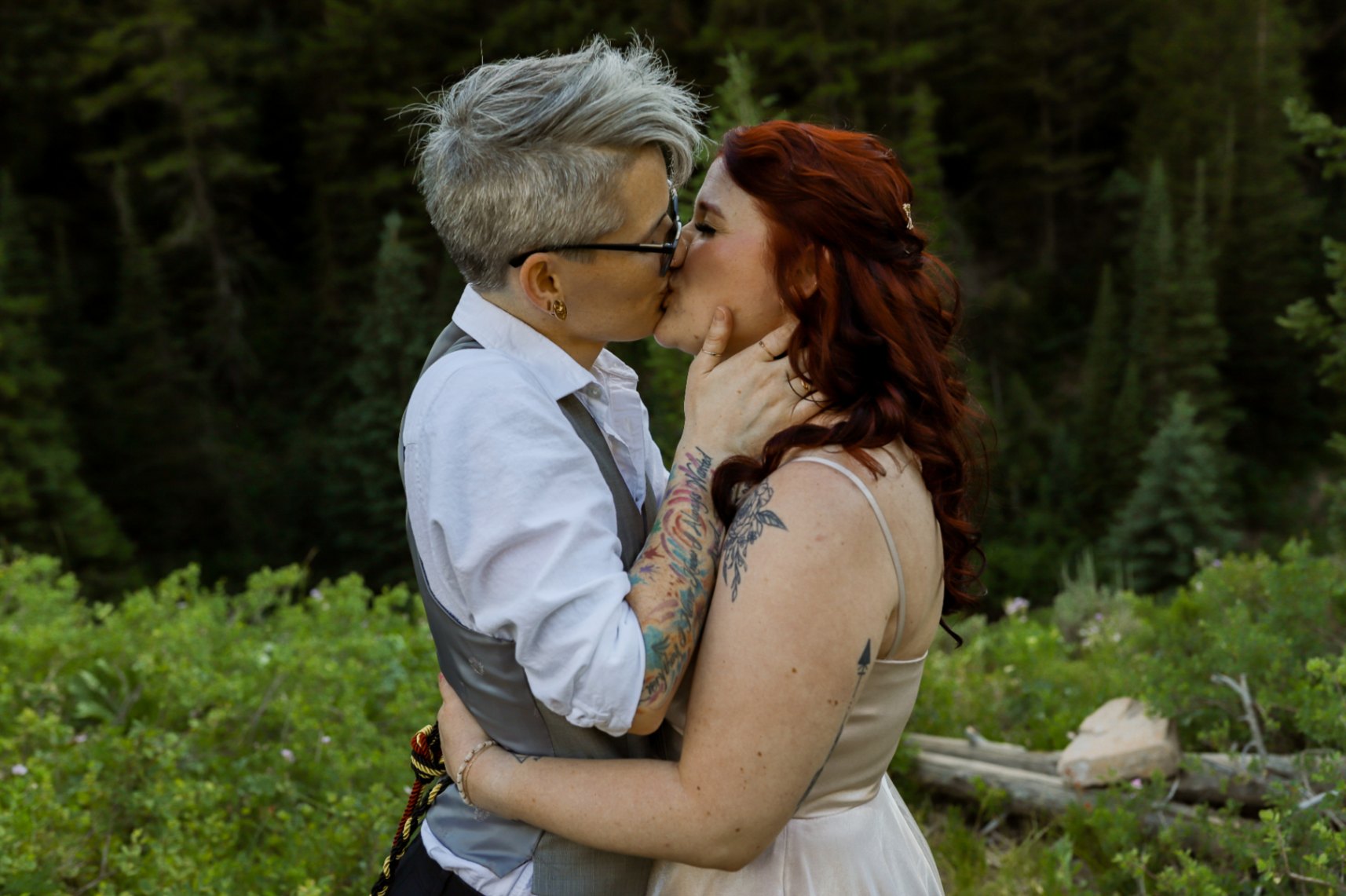 Two lesbian brides (both white, one with short gray hair, one with long dark red hair) kiss during their handfasting ceremony. They each have tattoos. The nonbinary bride with short gray hair is wearing a gray vest, a white shirt and dark pants, as well as glasses. The red-haired bride is wearing a light pink dress with spaghetti straps.