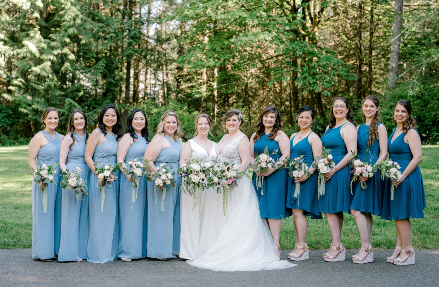 lesbian brides, one in a white wedding gown, and one in a white bridal jumpsuit, with their wedding party and bridesmaids, some in in teal blue knee length dresses and others in light blue pantsuits