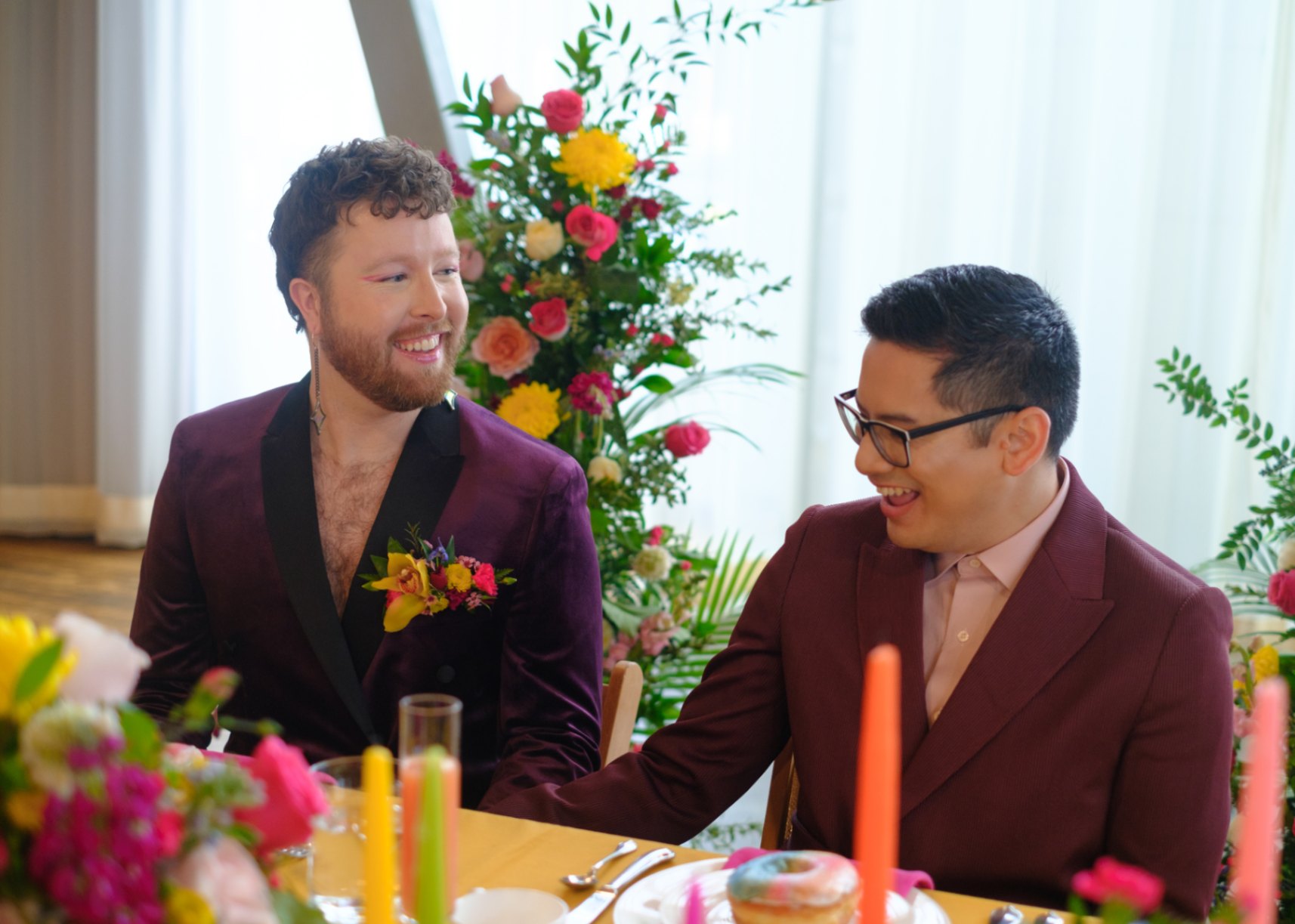 Colorful floral arrangements and a large disco ball hanging from ceiling for queer wedding inspiration while queer nonbinary couple chats and laughs. The linens and candles are yellow, green, orange and pink. The jackets are purple and burgundy.