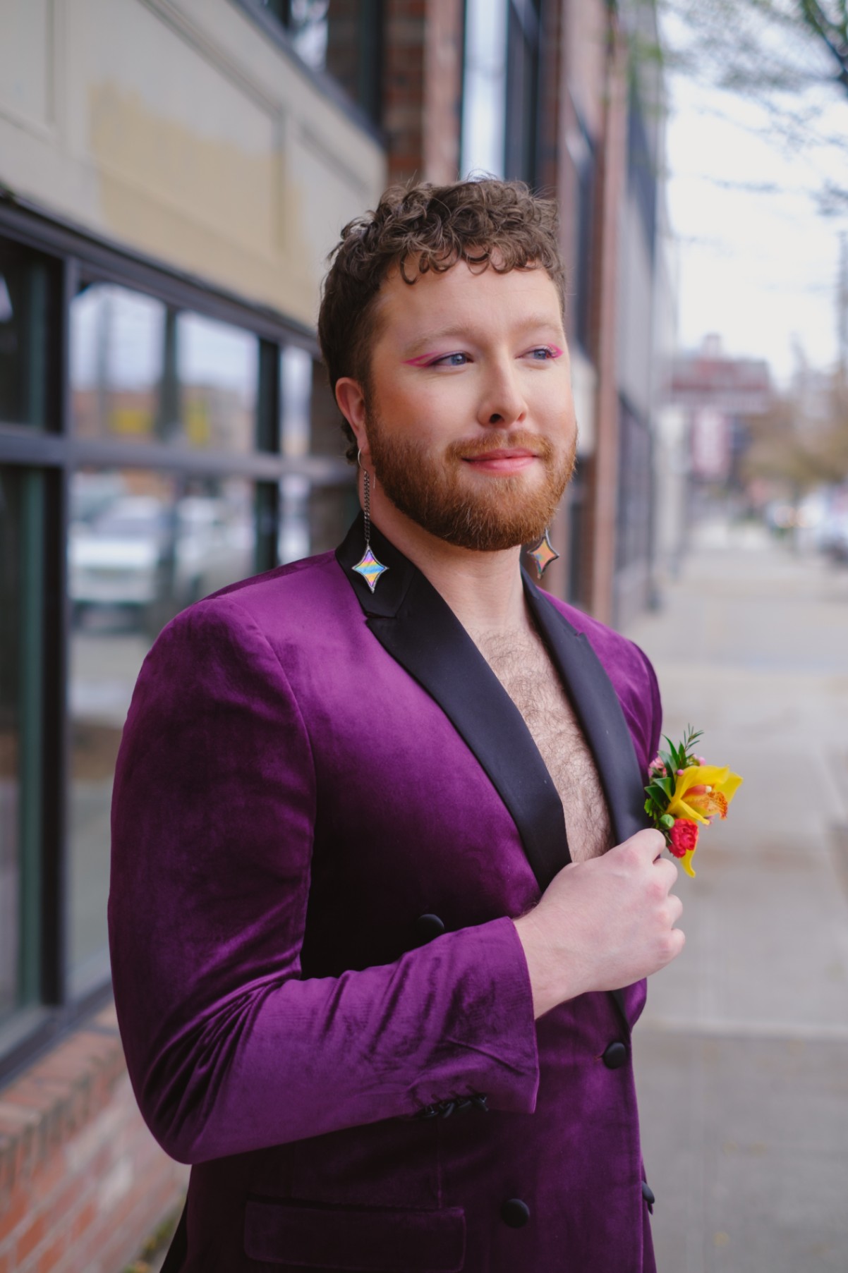 Nonbinary marrier with curly brown hair, a reddish-brown beard and hot pink eye liner gazes into the distance. They're wearing diamond drop earrings and wearing a velour purple tuxedo jacket with no shirt, showing off some chest hair.