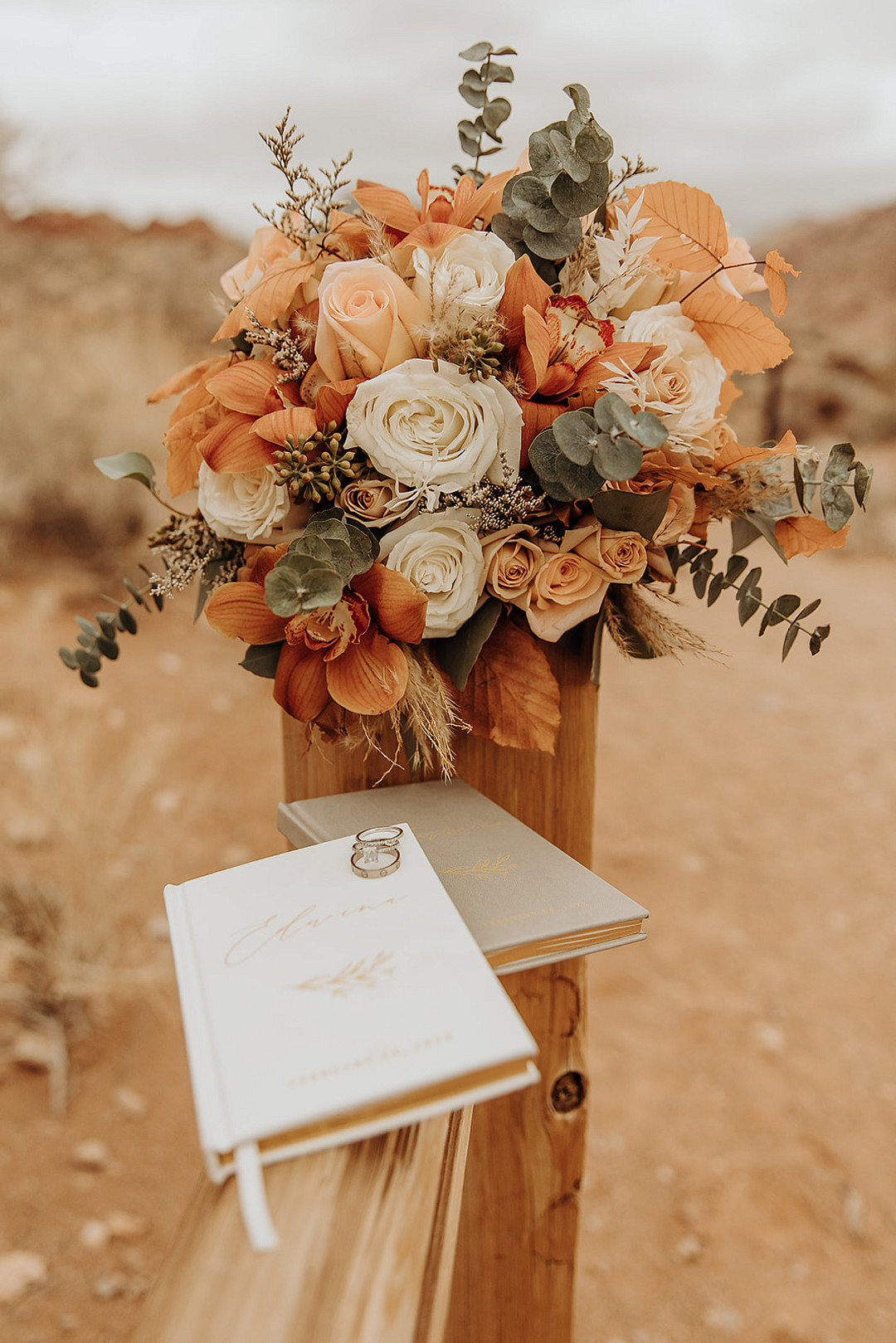 Wedding bouquet with white and peach roses, orange leaves, coral orange gladiolas, eucalyptus leaves and pampas grass. Also pictured: two brides vow books plus their wedding rings.