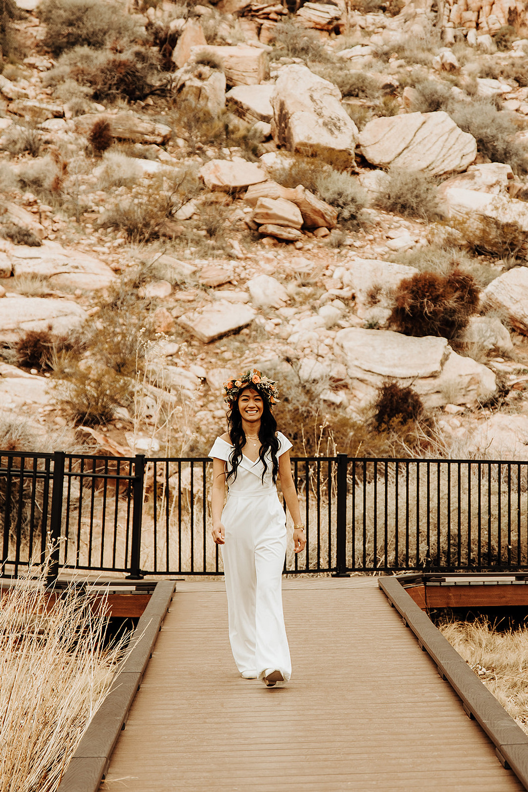 A bride with long dark hair walks up the aisle alone smiling. The bride is wearing an off-the-shoulder jumpsuit and a flower crown.