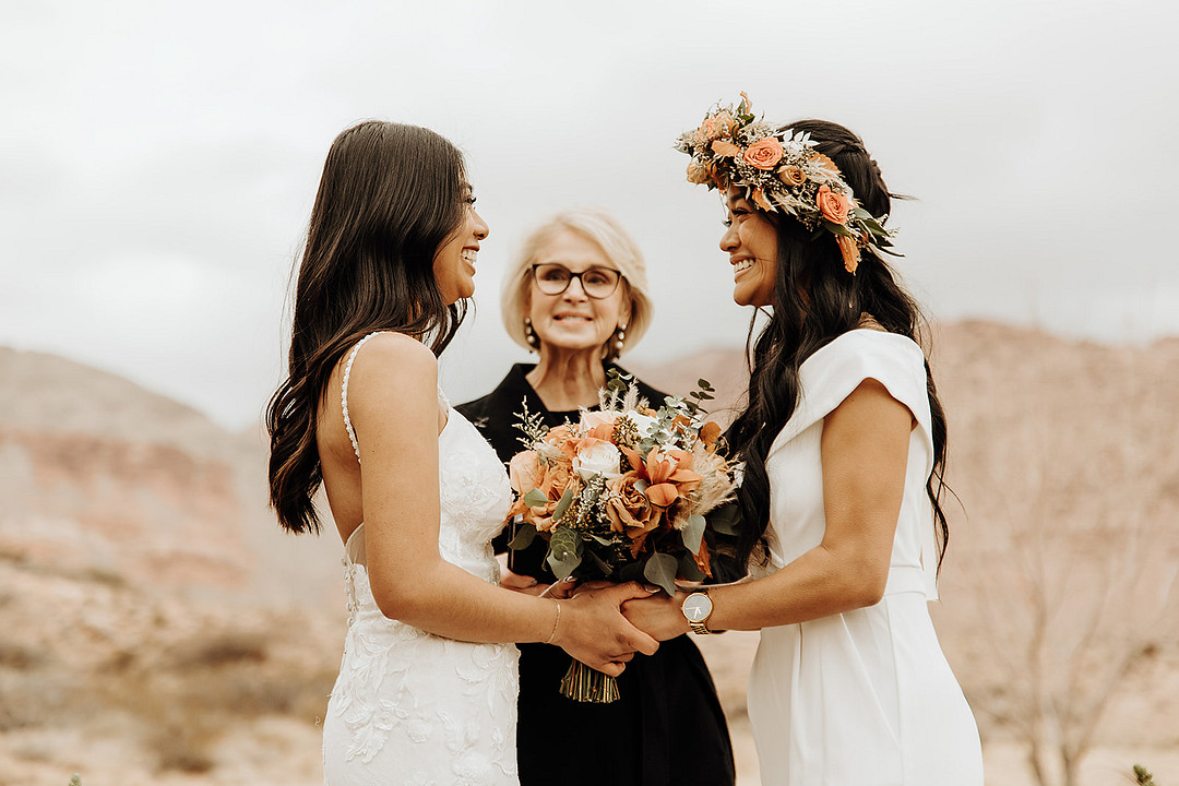 Two women of color exchange vows at their outdoor desert wedding. They both have long dark hair. In the center is the wedding officiant who is white with light blonde hair and dark glasses.