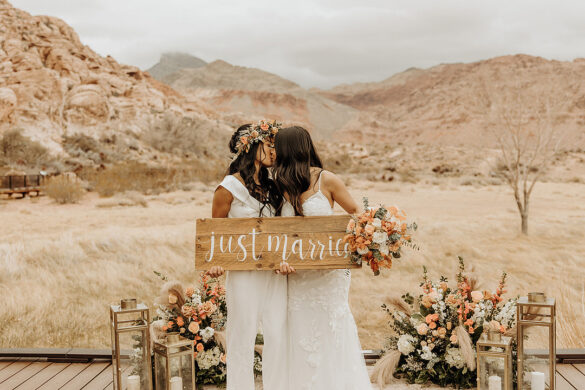 Two brides kiss while holding a Just Married wooden sign.