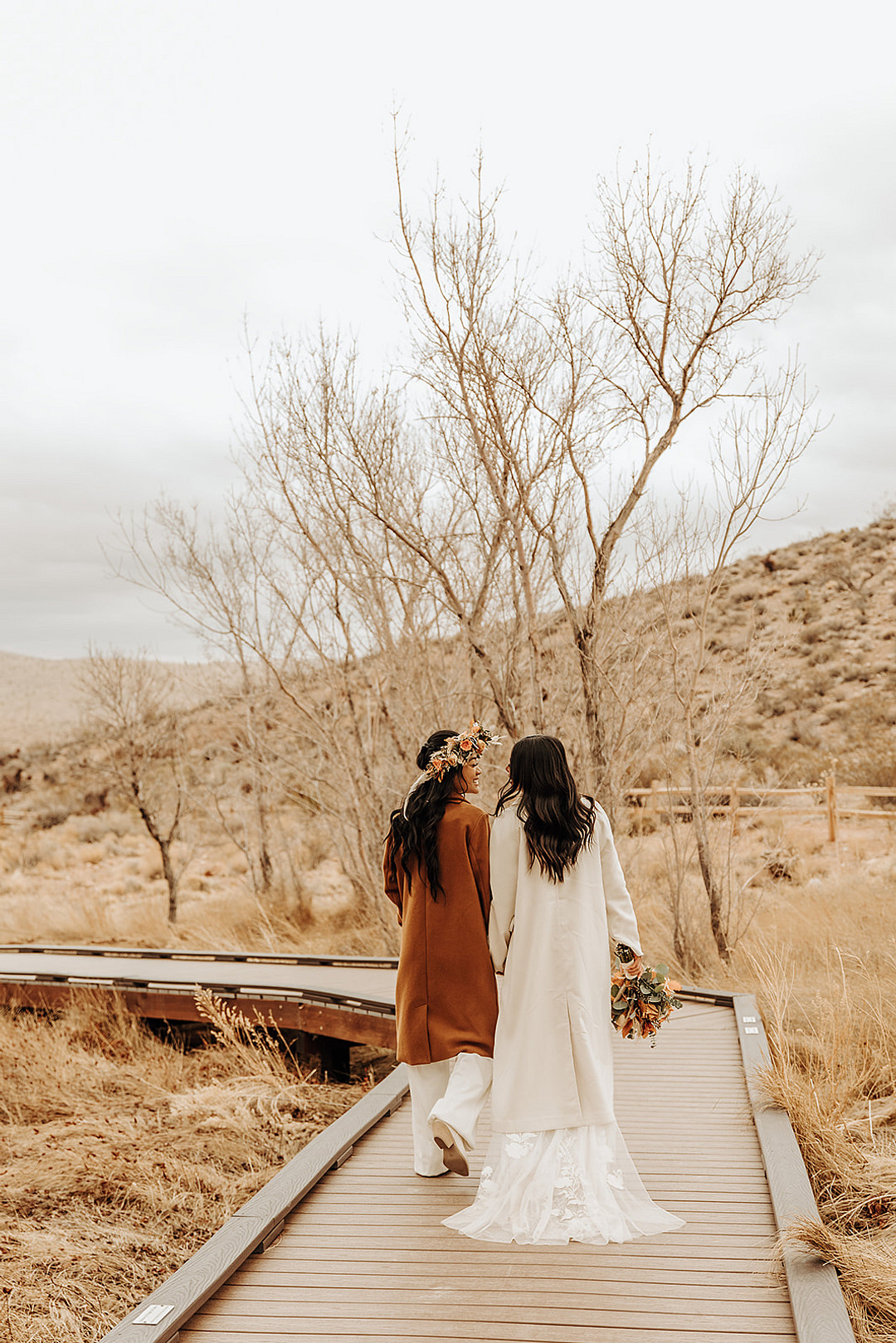 Two brides in wedding attire and overcoats walk on a boardwalk at their intimate Red Rock Canyon elopement.