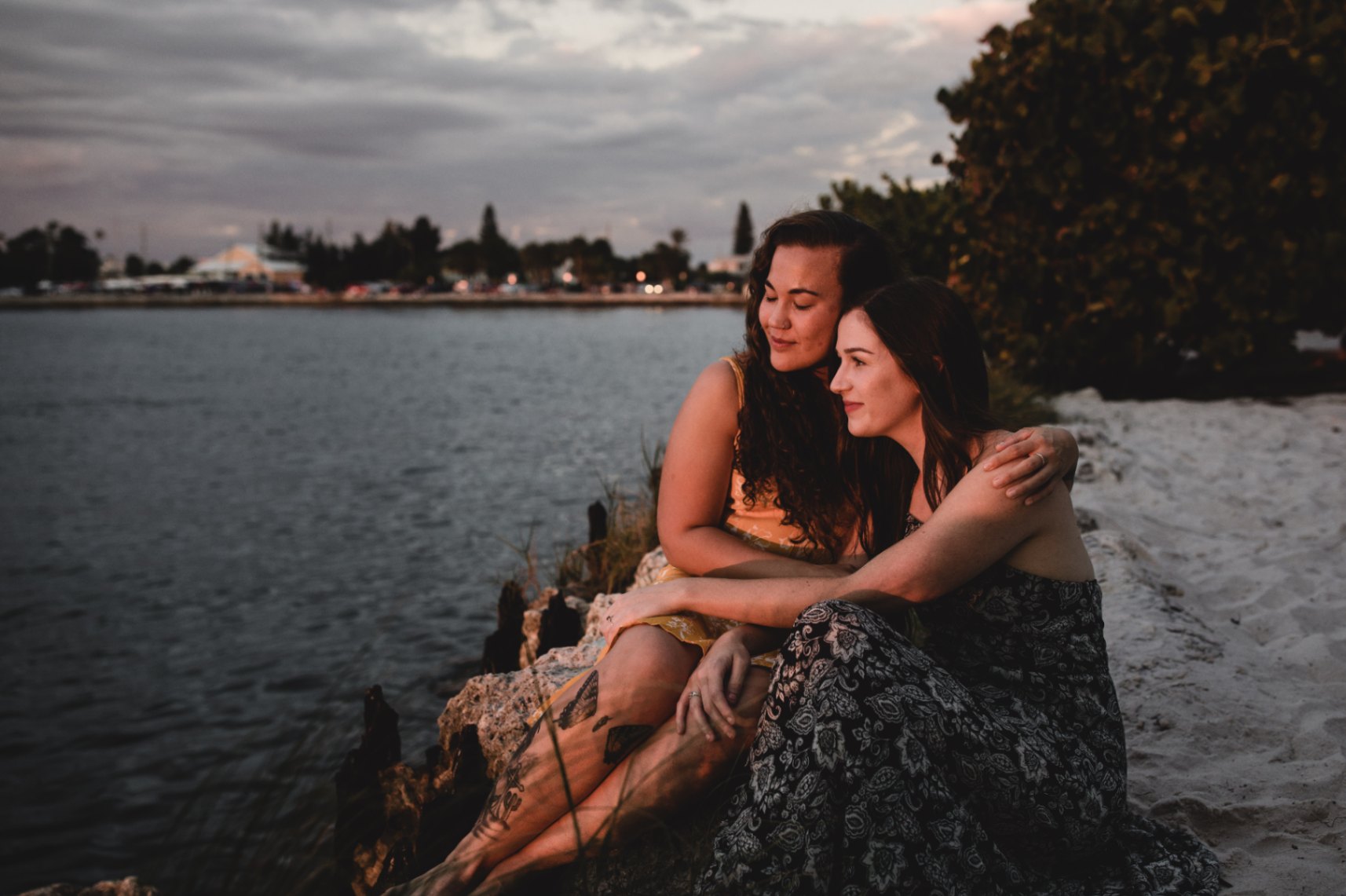 two women in love holding each other while sitting on beach and looking out at the sunset