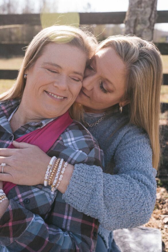 lesbians in love, women cuddling with a fence behind them. both are white and have straight blonde hair and are wearing brides beaded bracelets