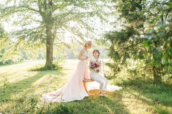 nonbinary lovers outside on their wedding day. they are both wearing light colored wedding attire and flower crowns. one is sitting in a wooden swing looking up at their beloved who stands beside them.