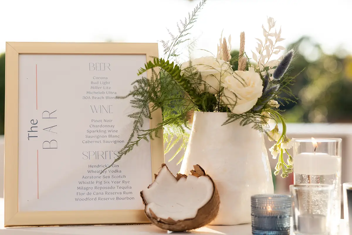 framed bar menu with beer, wine and spirits listed next to white vase of roses and natural plants, an open coconut, blue candle votive and clear candle votives