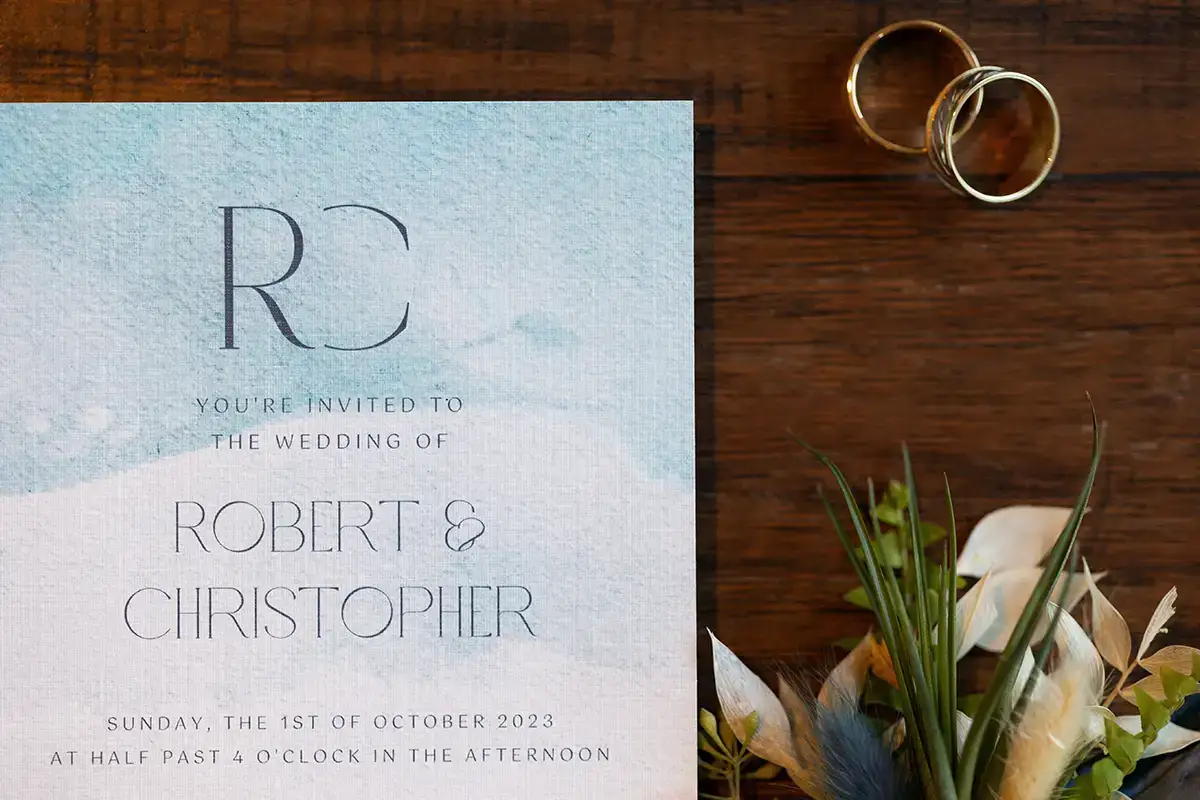 Blue watercolor invitation for LGBTQ+ wedding of two men flat lay with a boutonniere and two gold wedding rings