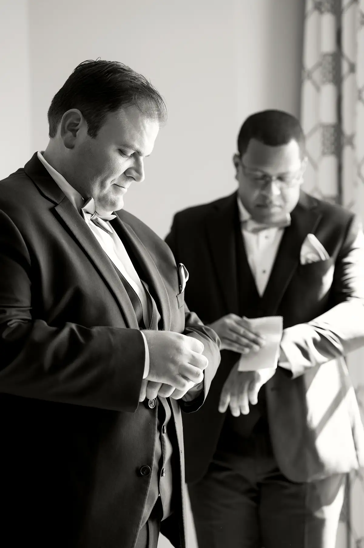 Black and white photo of two grooms getting ready together for their wedding. One groom is white, and the other groom is Black and wearing glasses.
