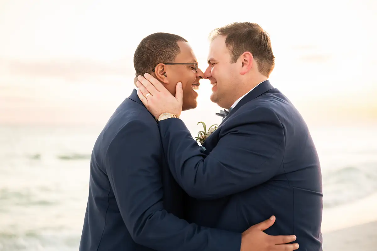 Interracial gay male couple smiling face to face with their arms around each other. The ocean is in the background. It is their wedding day.