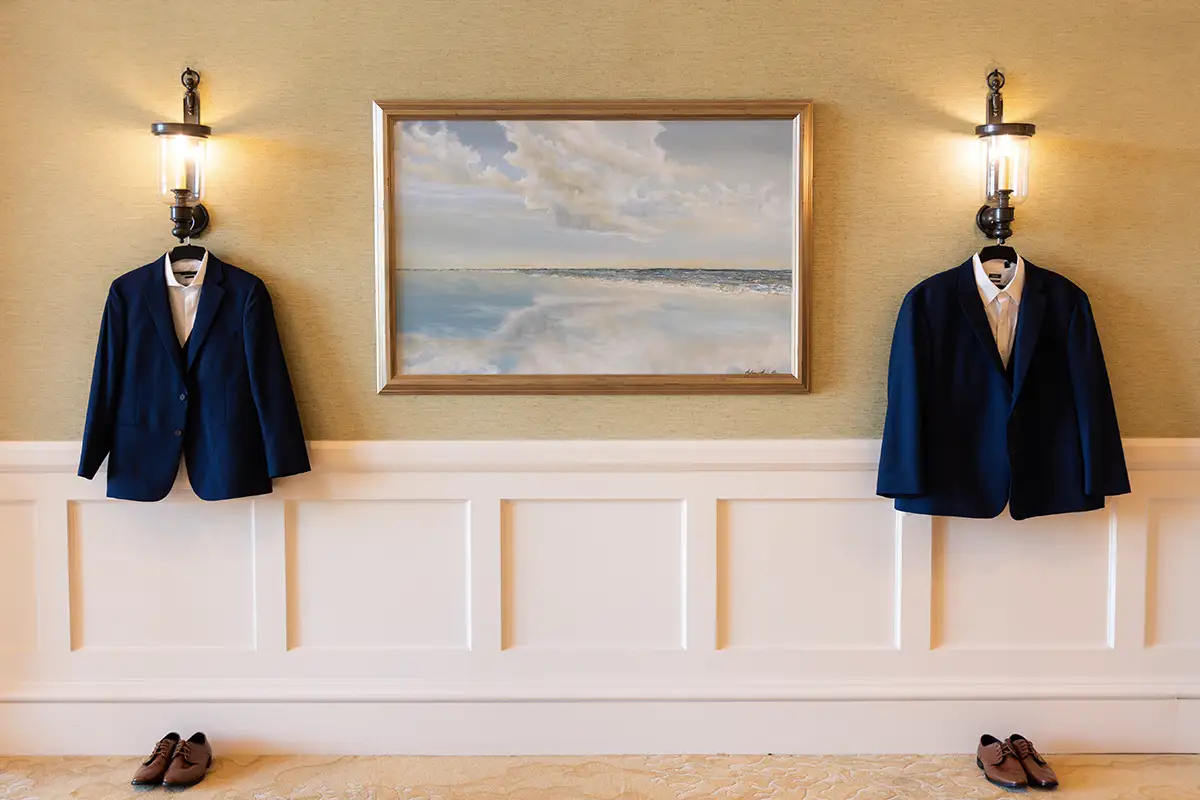 navy suit jackets hanging on golden wallpaper wall with ocean painting between them. brown shoes are on the floor