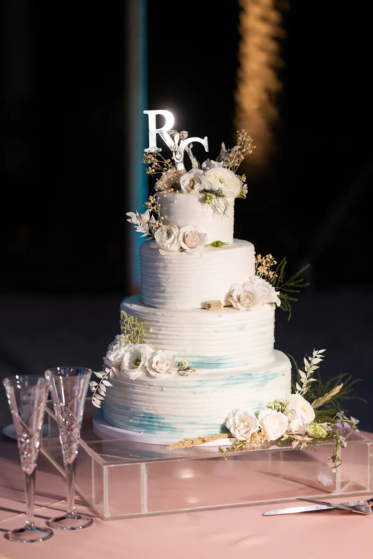 silver initials R and C on top of four-layer wedding cake with white and aqua blue frosting and white roses