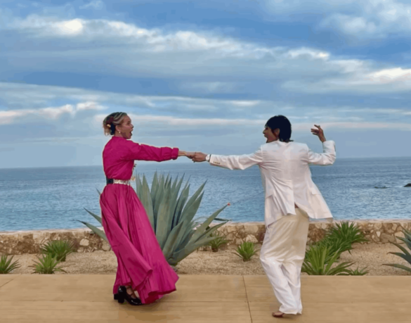Two women dance in front of the ocean. One is wearing a hot pink gown and the other woman is wearing a white suit.