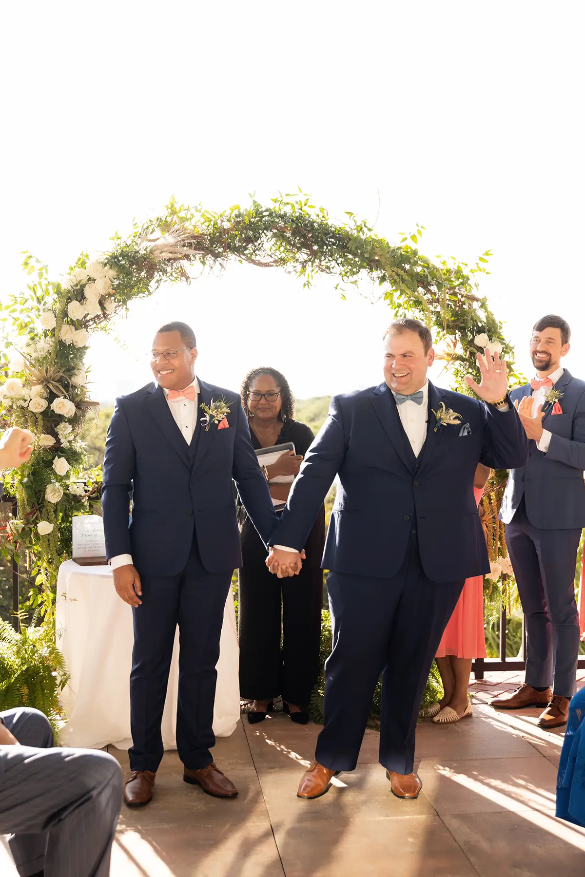 two-grooms-hold-hands-outdoor-coastal-gay-interacial-wedding. The Black groom is wearing glasses, a navy suit and a coral bow tie and pocket square. The white groom is wearing a navy suit, slate blue bow tie and pocket square.