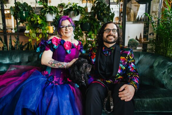 bride with colored hair in a colorful steampunk wedding gown and groom in a black suit with a rainbow floral pattern overlay and matching bow tie with their dog