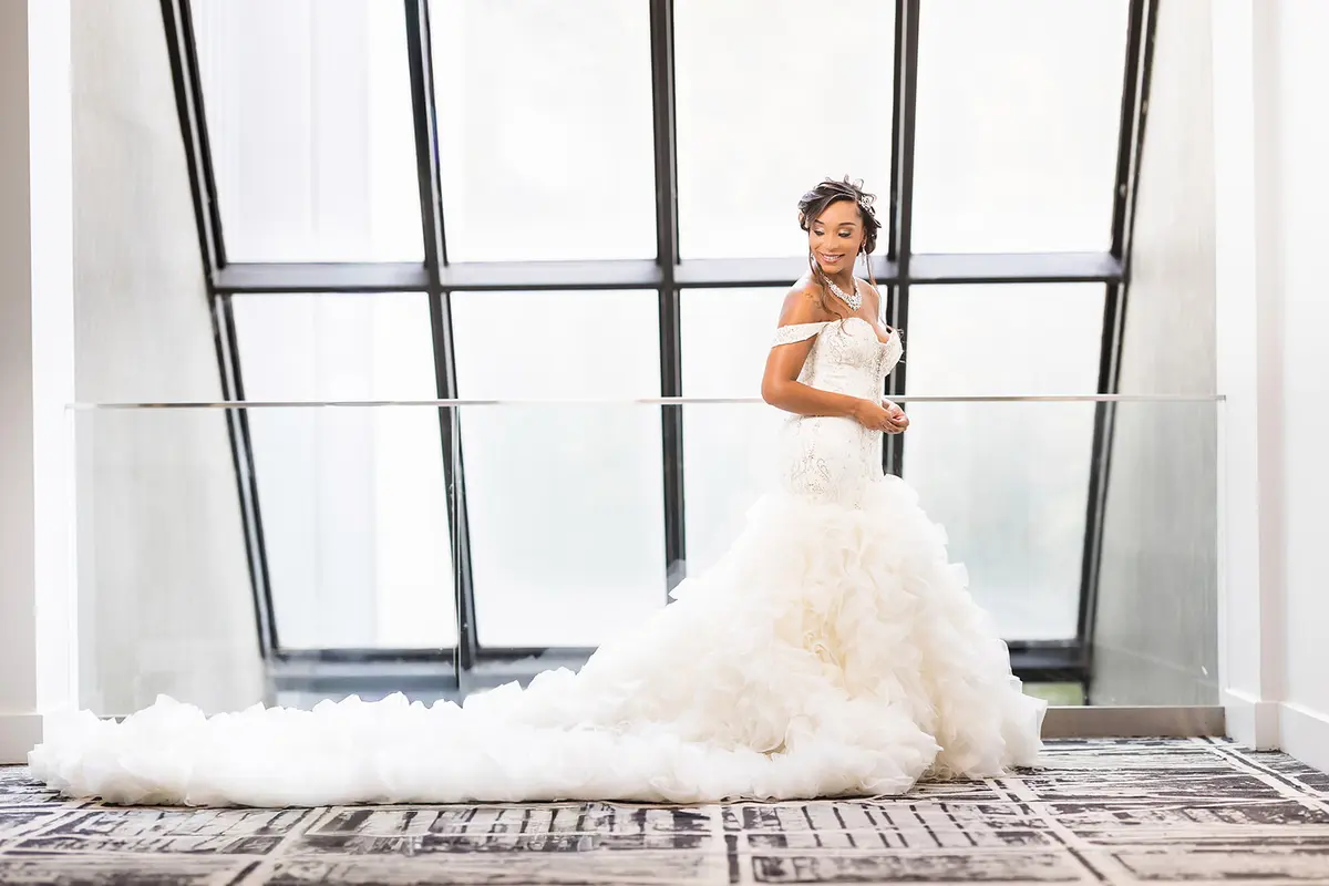 Black bride Daniella smiling in off-shoulder beaded mermaid wedding gown by window