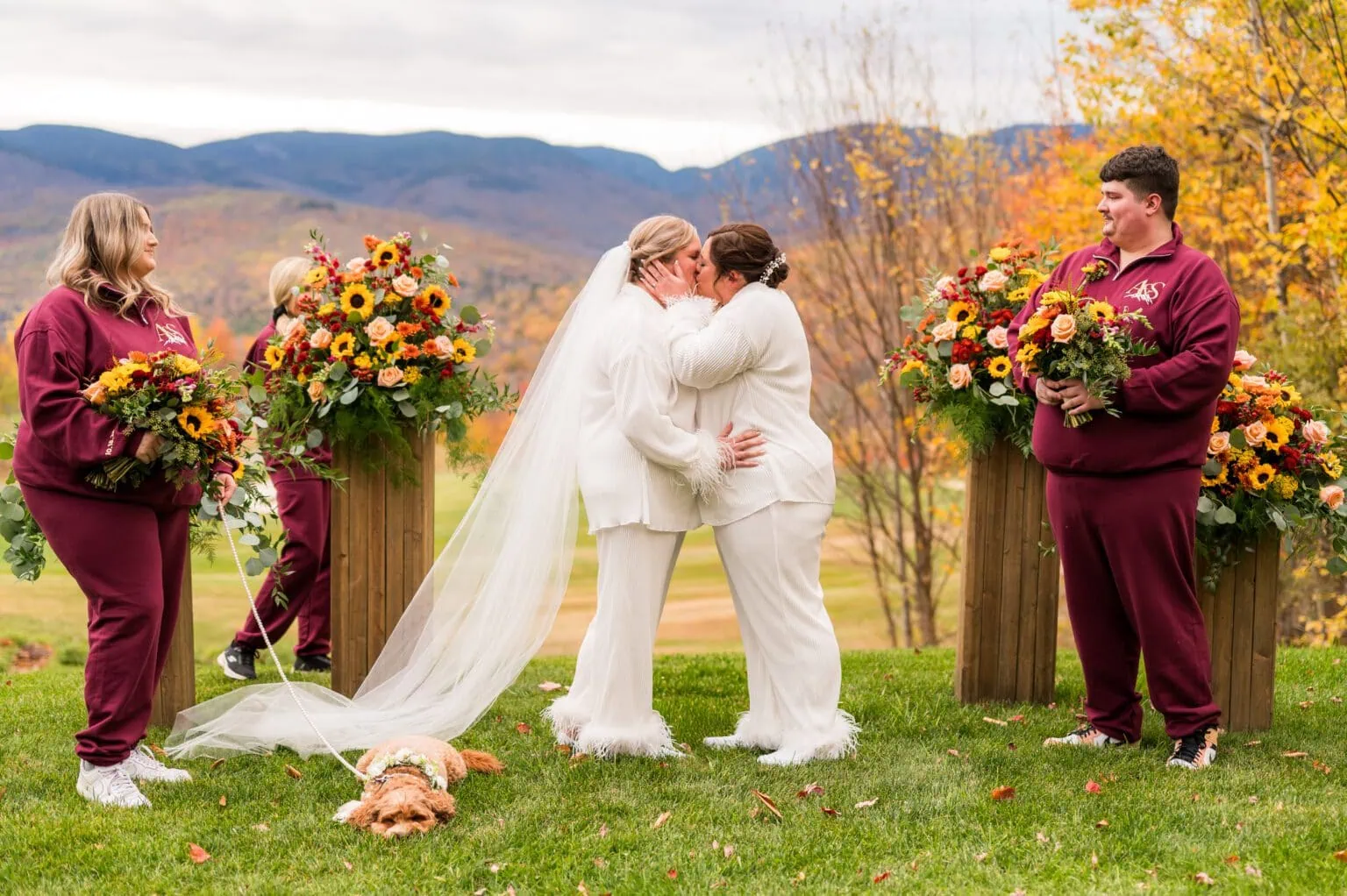 Two brides in bridal pajamas kissing while holding bouquets and their dog lying on the ground at their wedding party. Mountains are in the background.