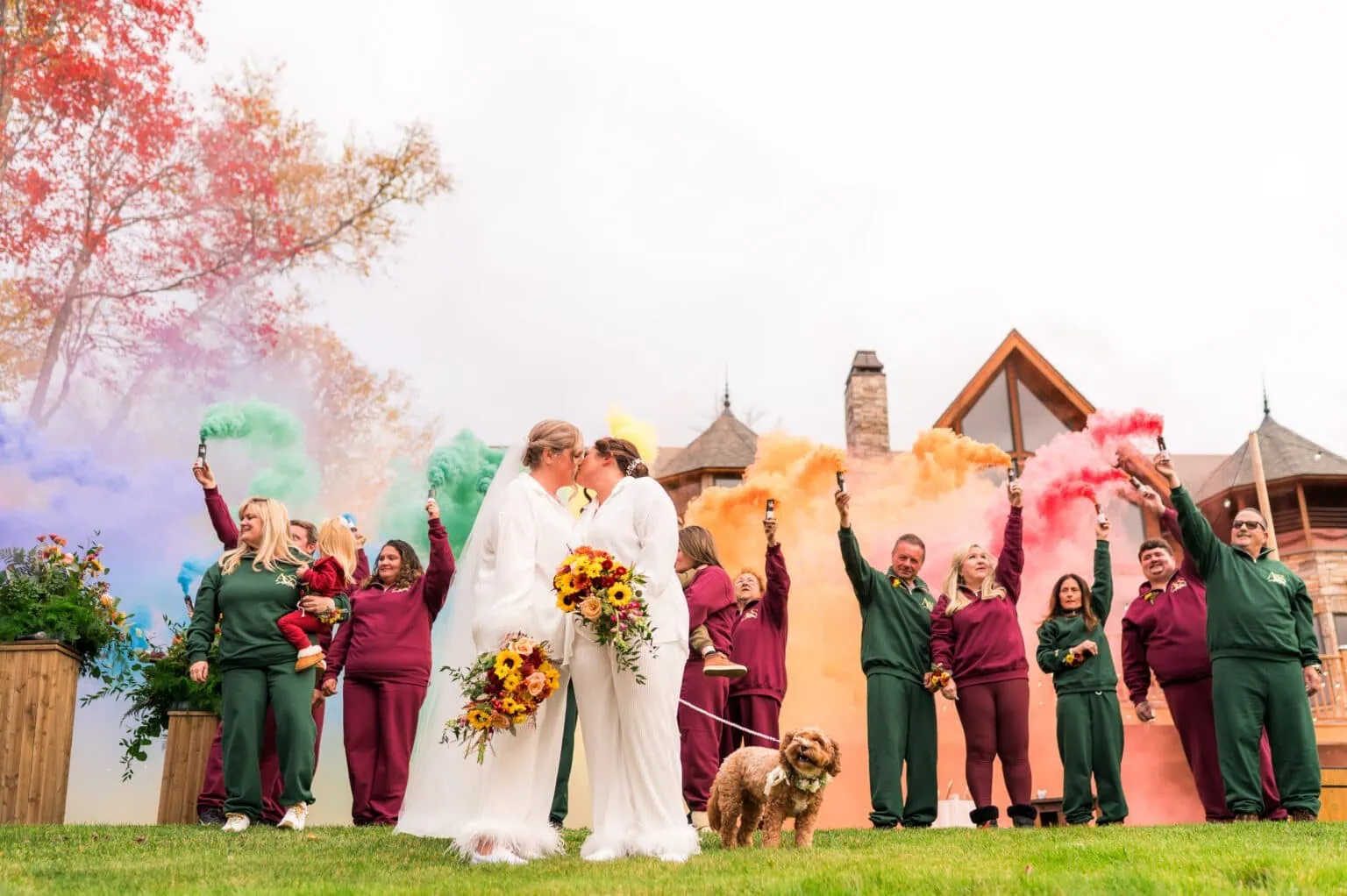 Two brides kissing while their wedding party holds rainbow-colored smoke bombs in the background