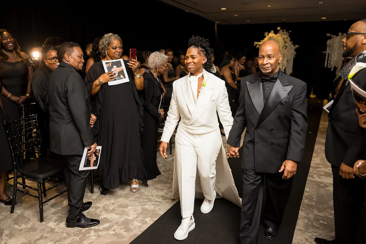 Black bride Tanaine in white suit walking down aisle with uncle at Atlanta lesbian wedding ceremony