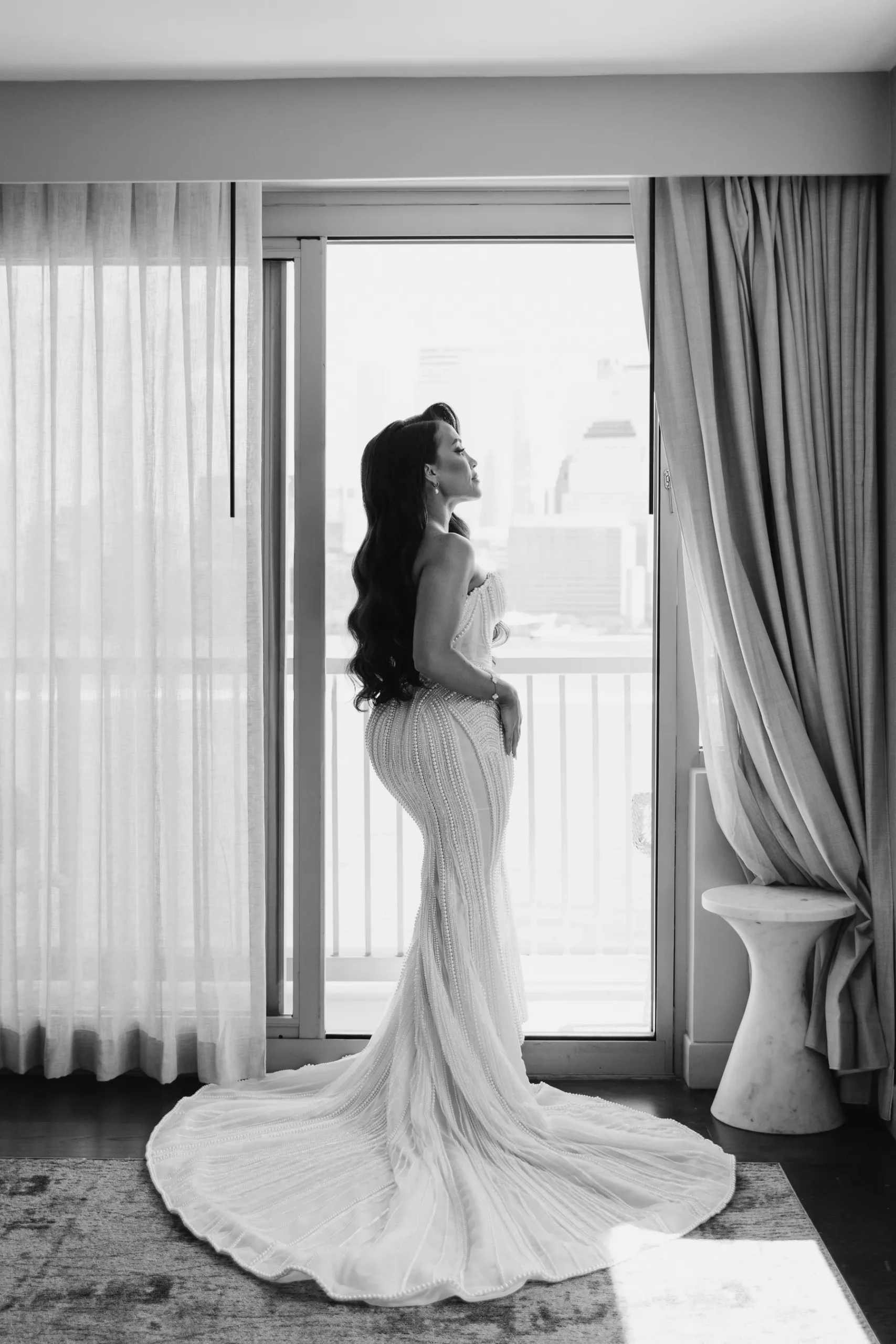 Bride in a pearl-beaded mermaid gown stands in profile at a sunlit balcony door at Hudson House in Jersey City.