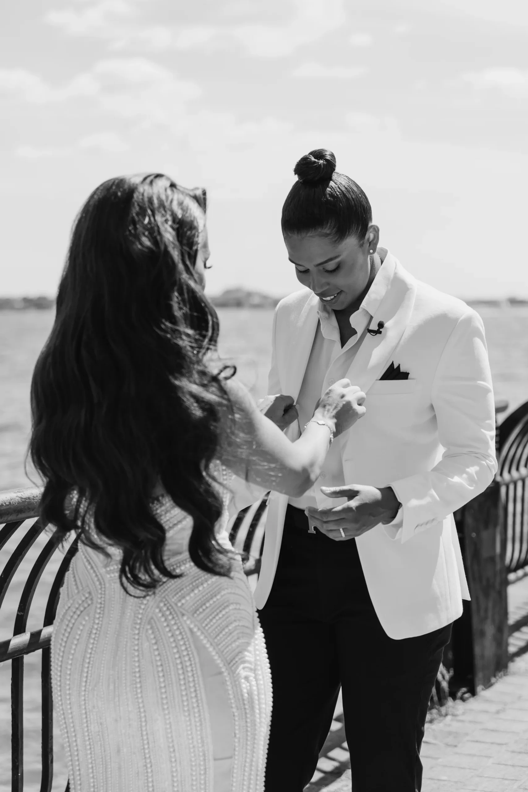 Michelle adjusts Katherine's white blazer cufflinks on a waterfront dock at Hudson House in Jersey City.