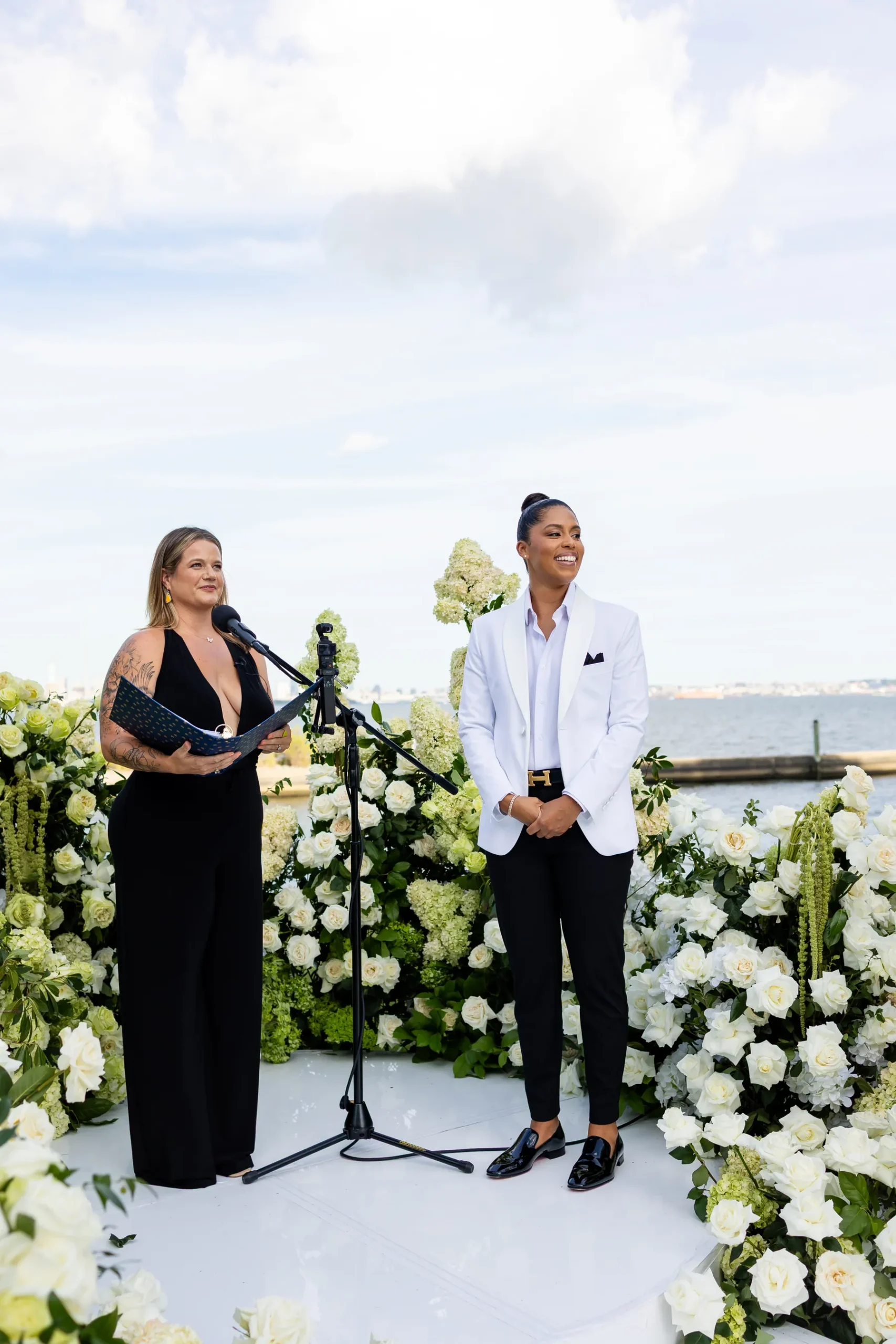 A person in a white blazer and black pants smiles at a floral-lined ceremony altar at Hudson House in Jersey City.