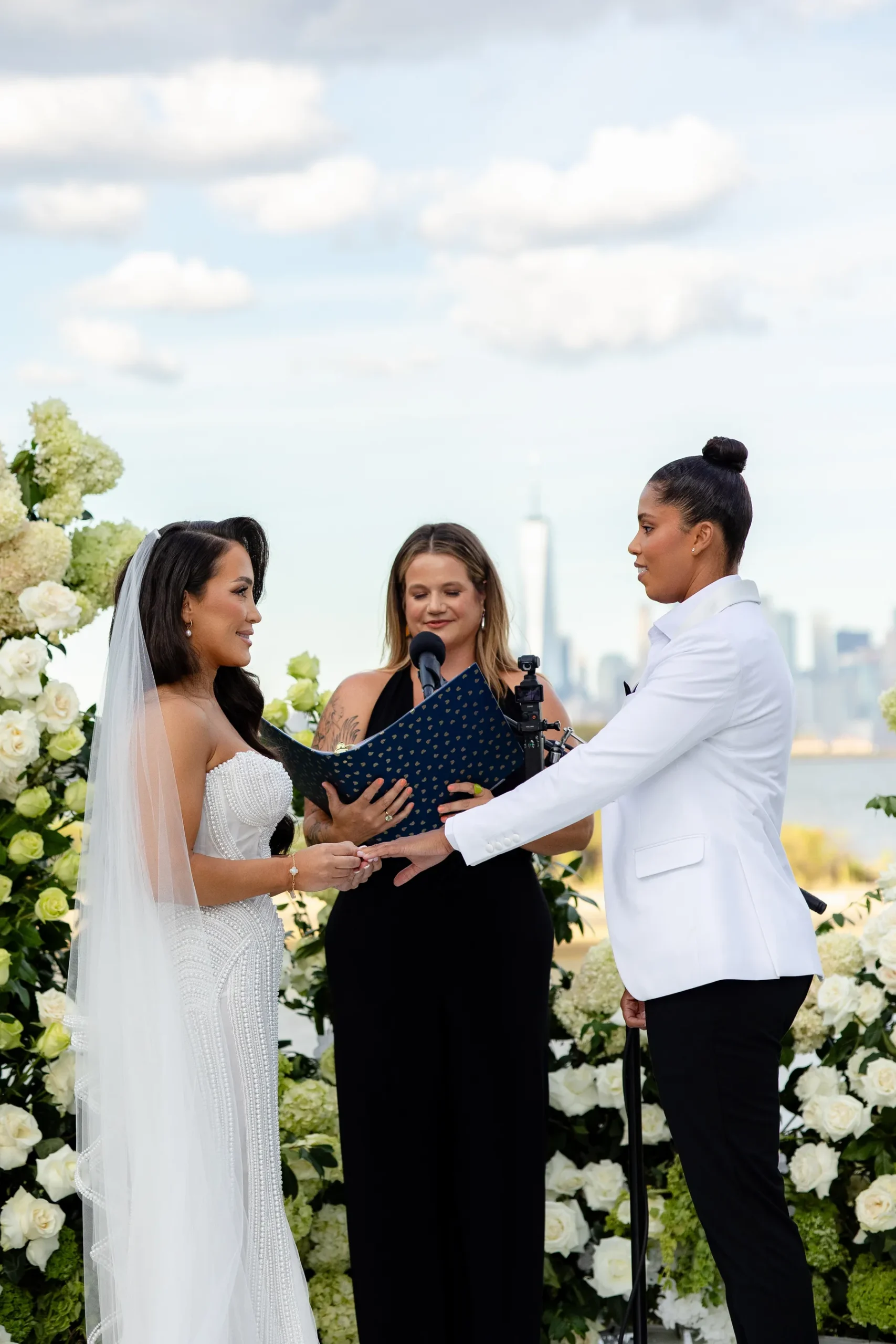 Katherine and Michelle exchange rings during their outdoor wedding ceremony at Hudson House in Jersey City, N.J., with the NYC skyline behind them.