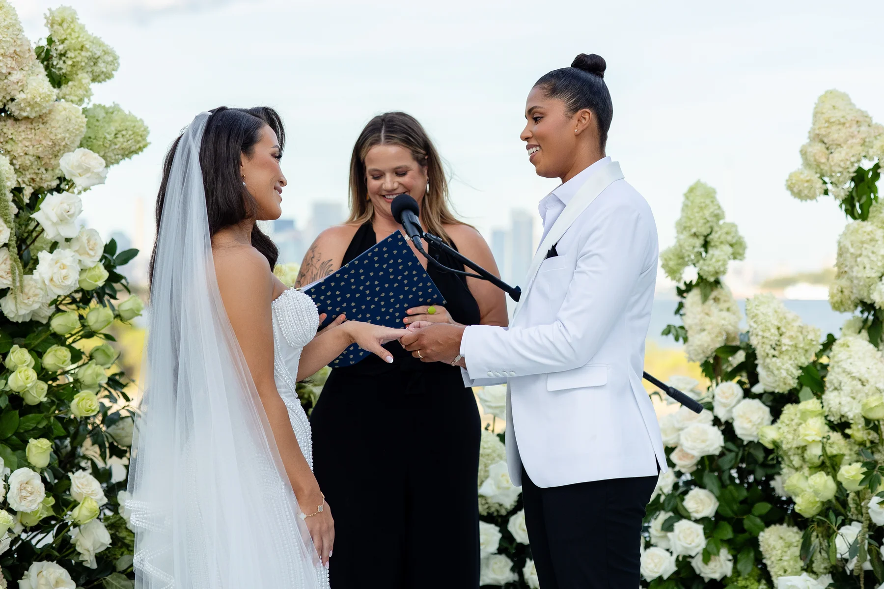 Katherine and Michelle exchange rings during their outdoor wedding ceremony at Hudson House in Jersey City, NJ, surrounded by white florals.