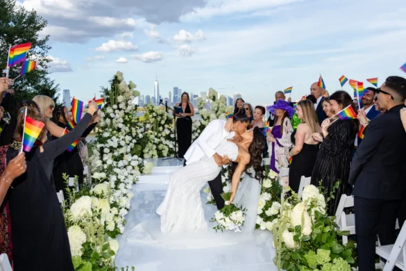 Katherine and Michelle share a dip kiss at their Hudson House wedding as guests wave rainbow Pride flags with the NYC skyline behind them.