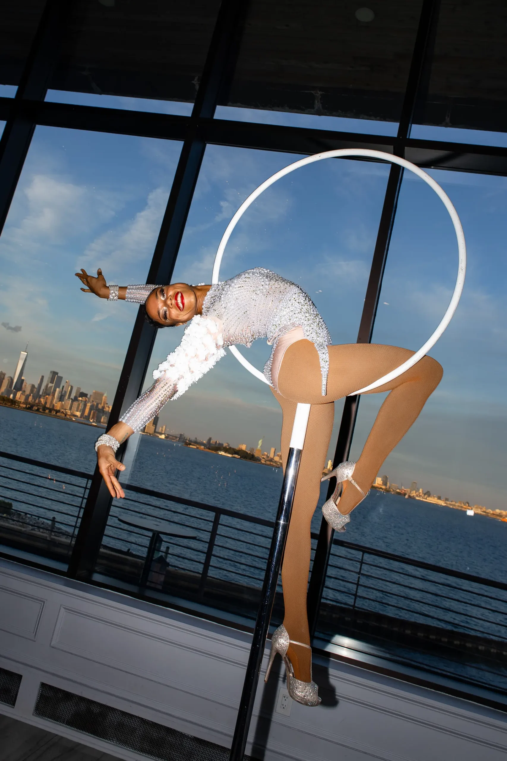 A bejeweled hula hoop performer bends dramatically at Hudson House in Jersey City with the NYC skyline behind her.