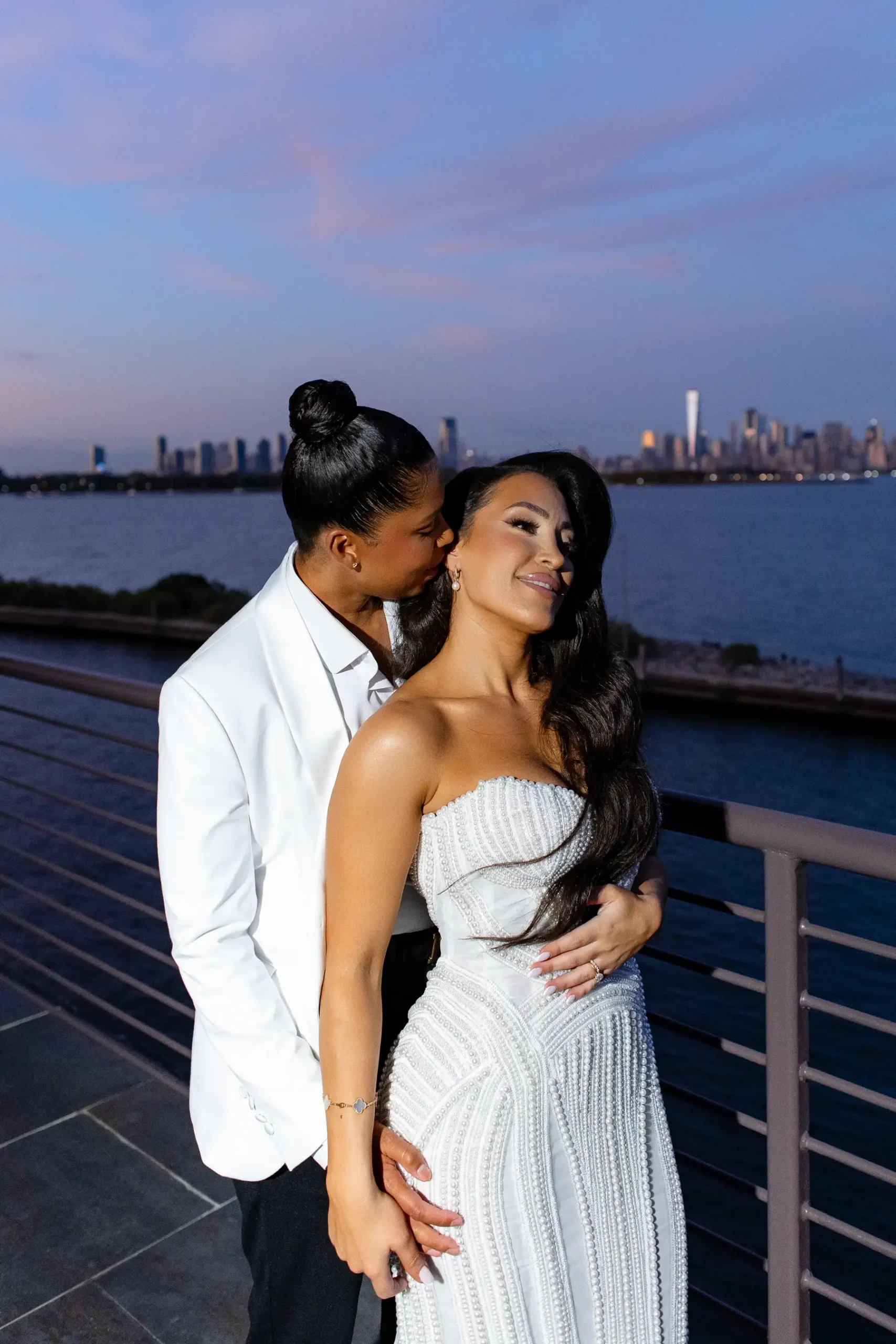 Katherine and Michelle share a romantic twilight embrace on the waterfront terrace at Hudson House in Jersey City with the NYC skyline behind them.