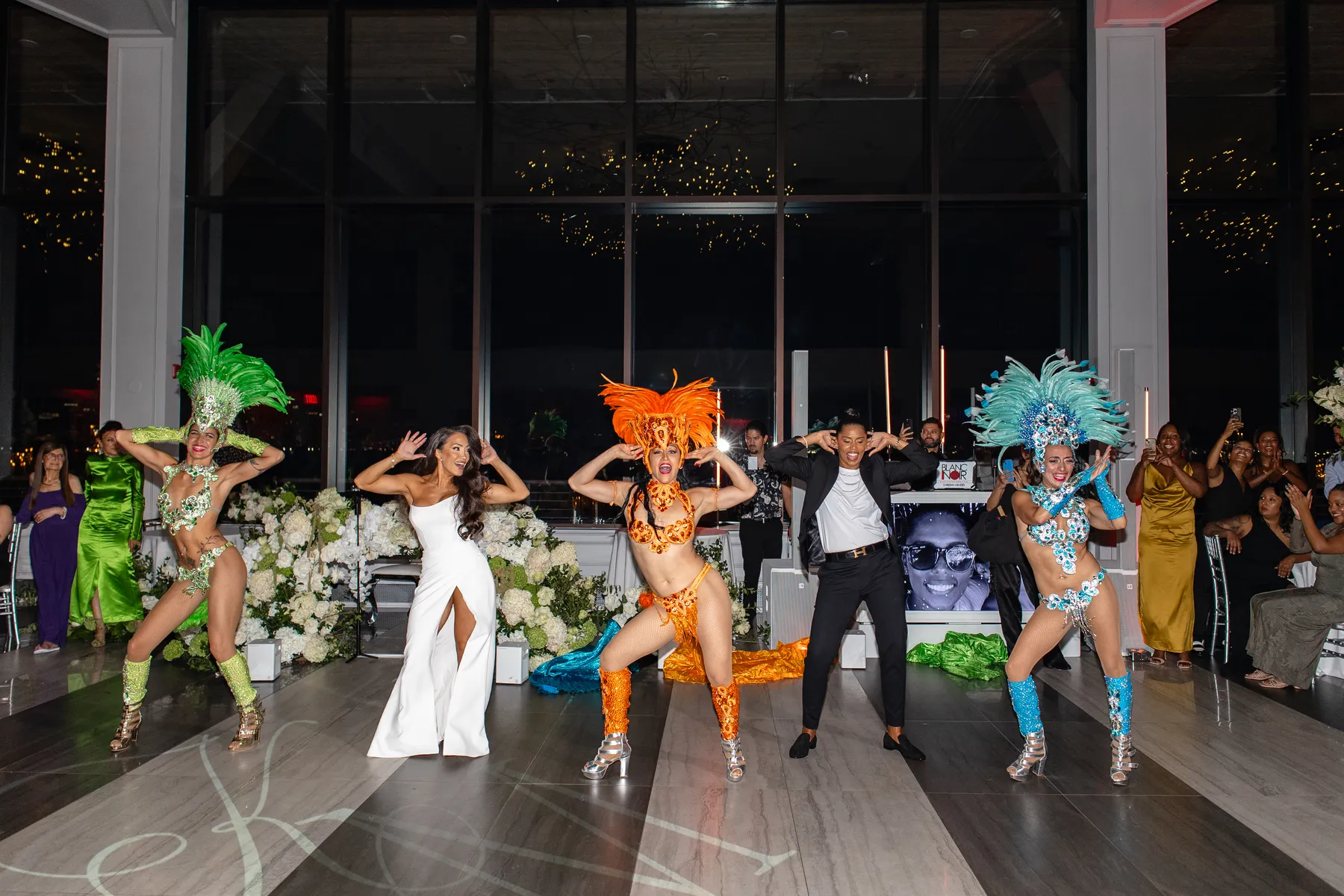 Katherine and Michelle dance with samba performers in feathered costumes at their Hudson House wedding reception in Jersey City.
