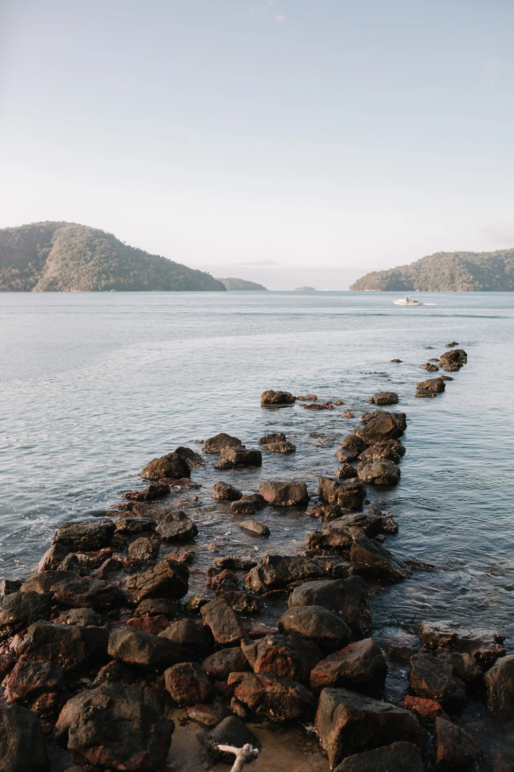 Rocky coastal shoreline with calm blue waters and green hills at Pouso das Flores in Paraty, Brazil.