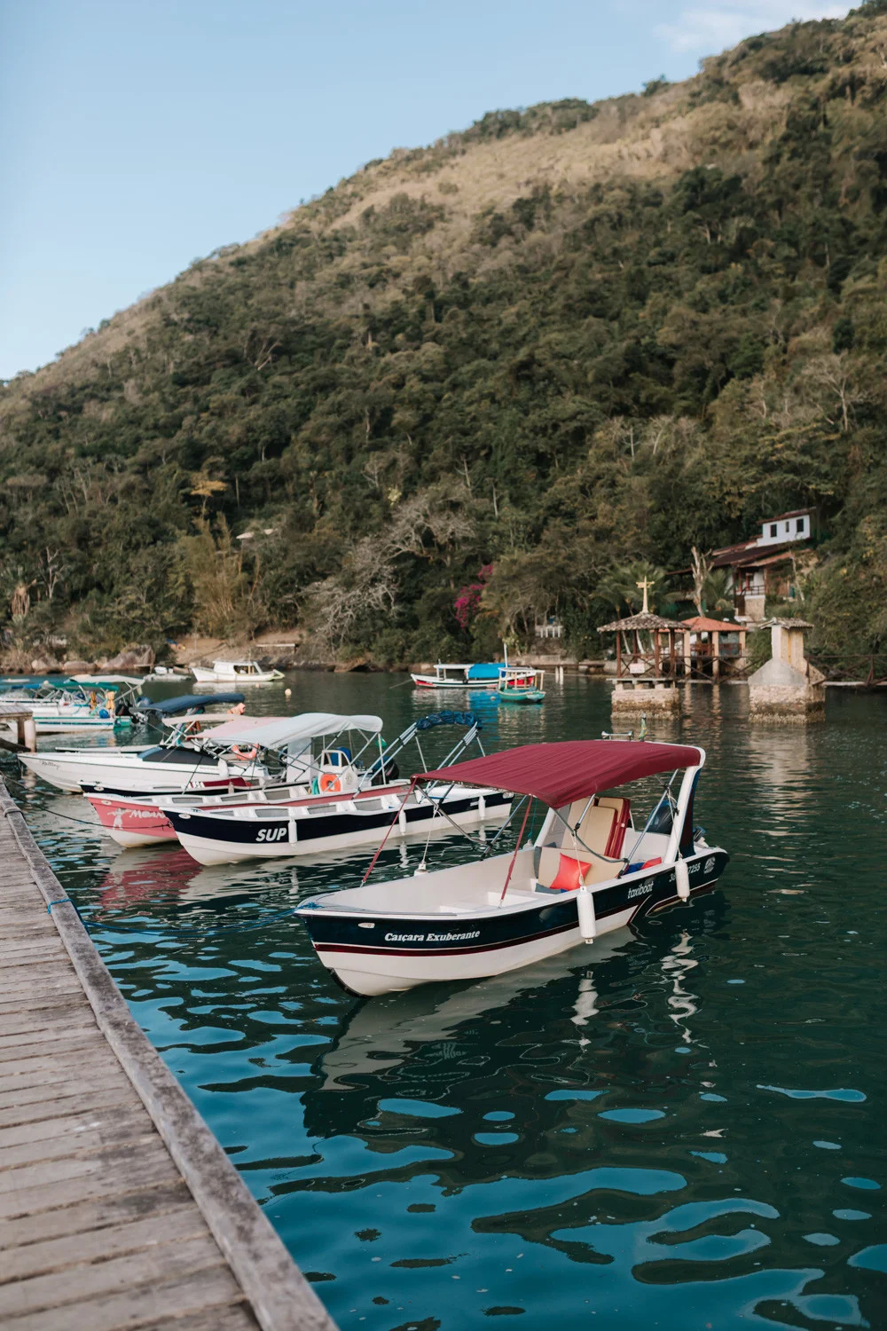 Small boats docked at a wooden pier in Paraty, Brazil, with lush green hills in the background at Pouso das Flores.