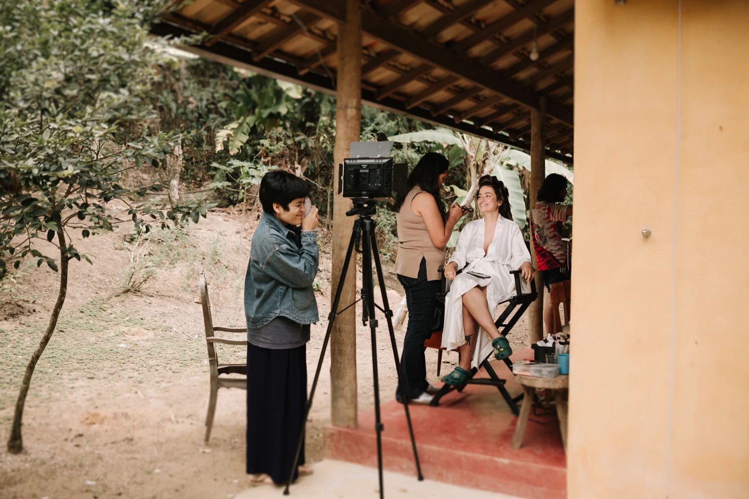 Bride Rafaela smiles in a white robe as a makeup artist works on her at Pouso das Flores in Paraty, Brazil.
