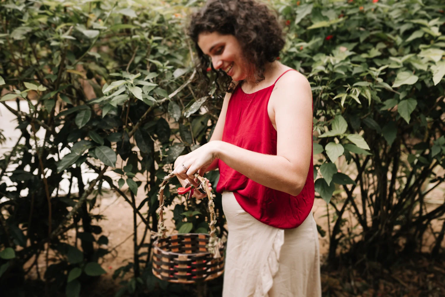 A smiling woman in a red top holds a woven basket while picking peppers in a lush garden at Pouso das Flores.