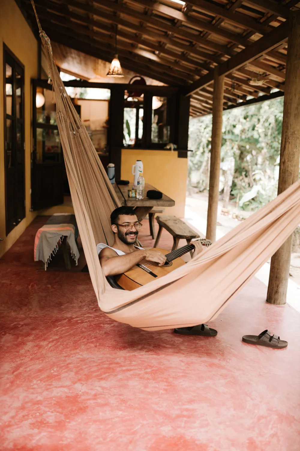 A smiling man plays acoustic guitar while relaxing in a hammock on the porch at Pouso das Flores in Paraty, Brazil.