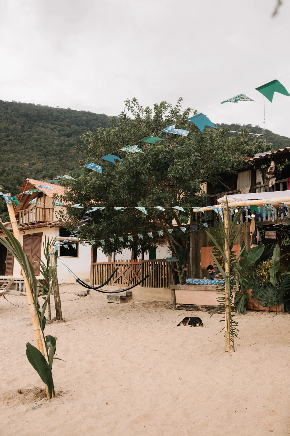 Sandy beachfront courtyard at Pouso das Flores in Paraty, Brazil, with hammocks, bunting flags, and tropical greenery.
