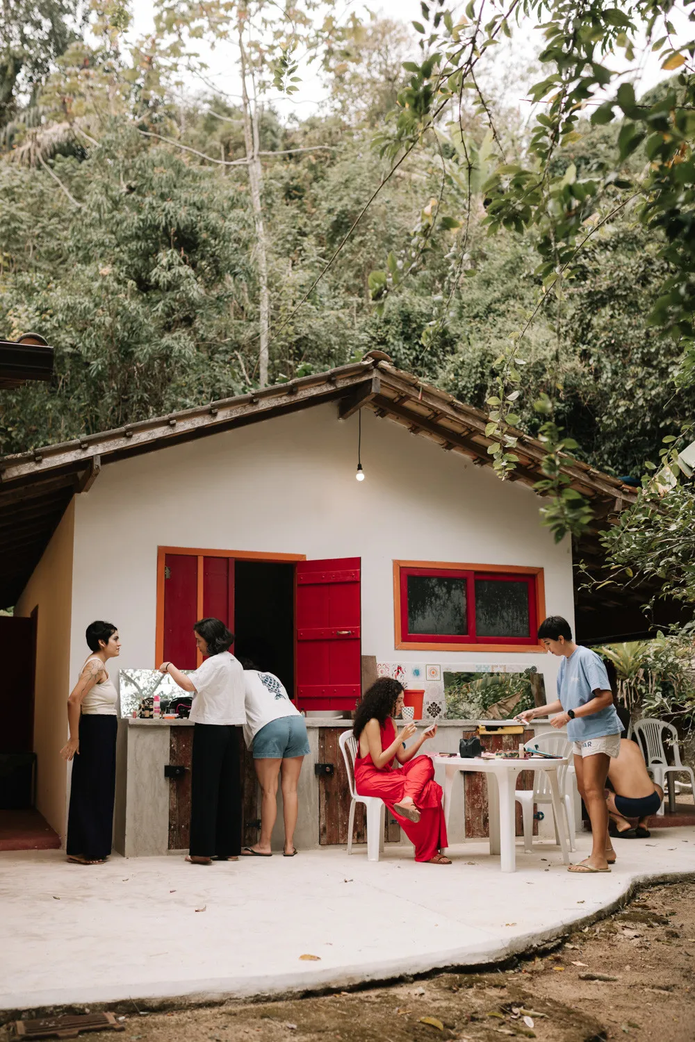 Guests gather outside a white cottage with red shutters at Pouso das Flores in Paraty, Brazil, during Rafaela and Nathalia's wedding.