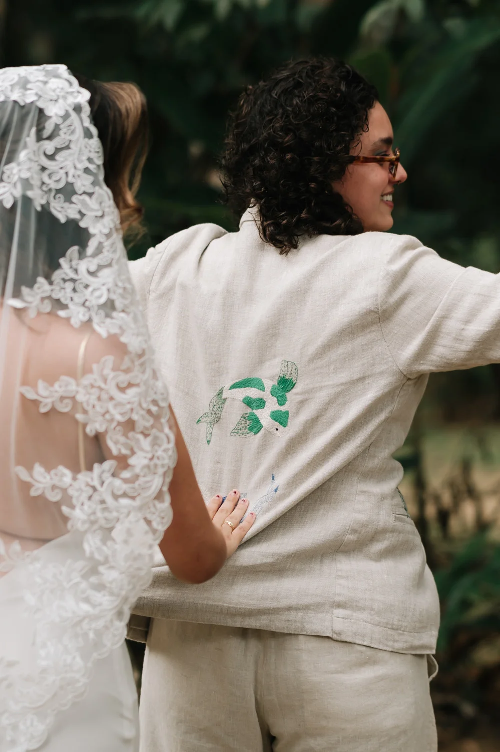Nathalia in a linen blazer with bird embroidery links arms with bride Rafaela in a lace veil at Pouso das Flores.