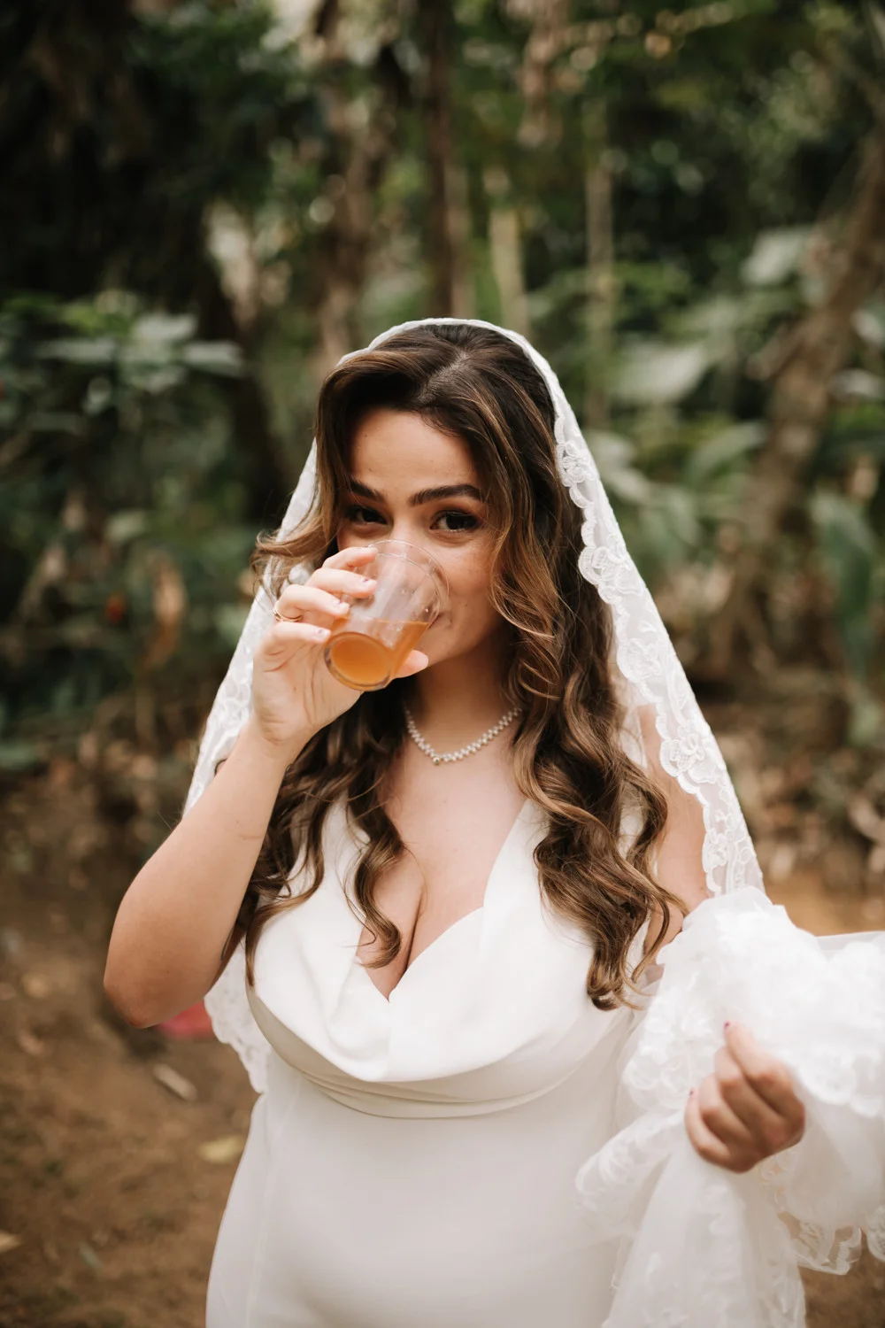Bride Rafaela in a white gown and lace veil sips a drink outdoors at Pouso das Flores in Paraty, Brazil.