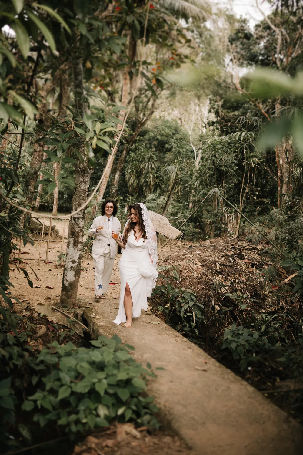 Rafaela and Nathalia walk a jungle path at Pouso das Flores in Paraty, Brazil, holding drinks and smiling.