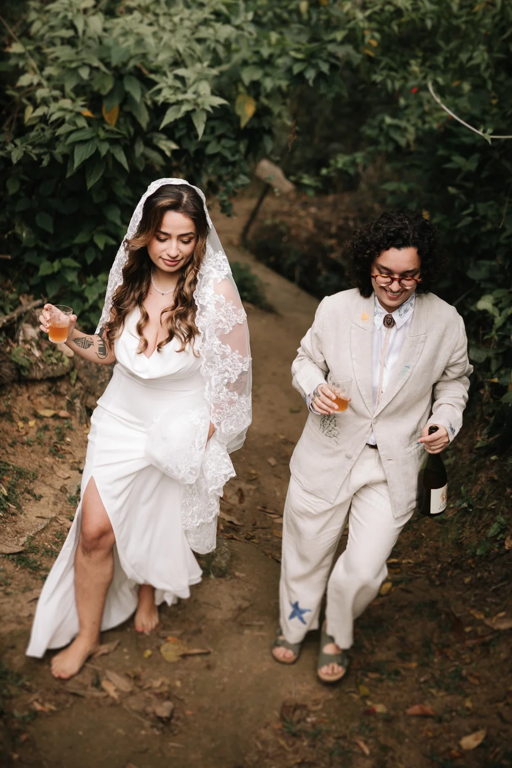 Rafaela and Nathalia walk a forest path at Pouso das Flores in Paraty, Brazil, laughing and holding drinks.
